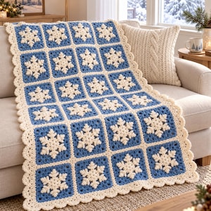 May include: A rectangular crochet blanket featuring a grid of blue square panels, each centered with a raised white snowflake motif. The blanket is finished with a scalloped cream-colored border and is constructed from yarn.