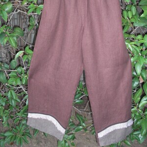 May include: Brown linen pants with an elastic waistband, hanging on a white hanger against a backdrop of green foliage. The hem of the pants features a contrasting gray fabric with a white decorative trim.