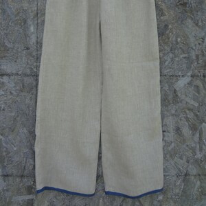 May include: Tan linen pants with a wide leg design, hanging on a white hanger. The pants have a blue trim along the bottom hem. The fabric appears to be lightweight and suitable for warm weather.