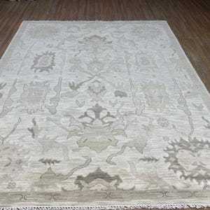 Könnte beinhalten: Ein rechteckiger Teppich mit hellcremefarbenem Hintergrund und einem sich wiederholenden Muster aus stilisierten Blumen- und geometrischen Motiven in Taupe-, Beige- und sanften Grautönen. Der Teppich hat kurze Fransen an den kurzen Seiten.