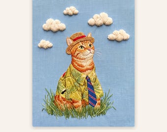 Op de afbeelding: Een borduurwerk op lichtblauwe stof met een oranje cyperse kat die een strooien hoed, een geel overhemd met patroon en een blauw-rode gestreepte das draagt. De kat zit in het groene gras onder vier witte geborduurde wolken.