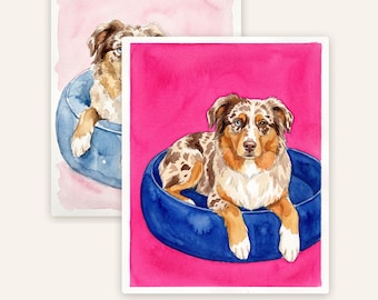 Op de afbeelding: Een aquarelillustratie van een bruin-witte Australian Shepherd hond met heterochromie, zittend in een ronde blauwe hondenmand tegen een levendige magentakleurige achtergrond. Een tweede, soortgelijke illustratie met een lichtroze achtergrond is zichtbaar.