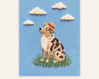 Op de afbeelding: Een borduurwerk op lichtblauwe stof met een bruin-witte gevlekte hond die een strooien hoed met een rode band draagt. De hond zit in groen gras onder een lucht met vier witte geborduurde wolken. De tekst onderaan luidt: Preview in 24h · Unlimited revisions.
