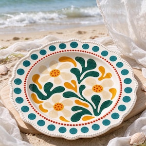 May include: A round ceramic plate with a scalloped edge featuring a cream-colored base. The rim is decorated with teal dots and a thin orange line. The center displays a floral pattern with cream flowers, orange centers, and dark green leaves.