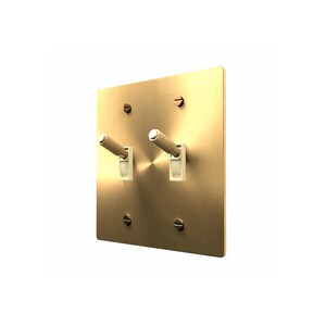 May include: A square, brushed brass light switch plate featuring two toggle switches with textured, knurled cylindrical handles. The plate is secured by four flat-head screws positioned at the corners, and the toggle mechanisms are set in white plastic.