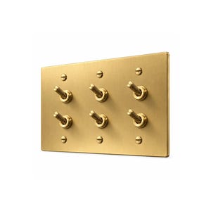 May include: A rectangular brushed brass light switch plate featuring six individual knurled toggle switches arranged in two rows of three. The plate is secured with three flat-head screws along the top and three along the bottom edge.