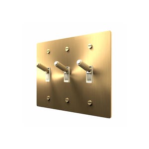 May include: A rectangular brushed brass light switch plate featuring three toggle switches with a textured, knurled grip. The plate is secured by six flat-head screws, with two screws positioned above and below each of the three individual toggle switches.