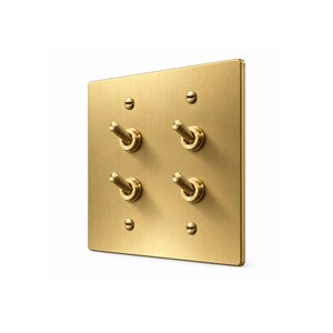 May include: A square, brushed brass light switch plate featuring four individual toggle switches arranged in a two-by-two grid. The plate is secured with four matching flat-head screws, and each toggle switch has a textured, cylindrical metal handle.