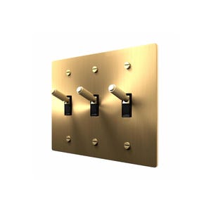 May include: A triple light switch wall plate with a brushed brass finish. The rectangular plate features three toggle switches with textured, knurled brass handles and black bases, secured to the wall with six flat-head brass screws.
