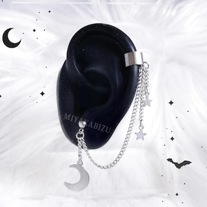 Könnte beinhalten: Silberner Ohrclip mit einer baumelnden Kette und einem Mond-Anhänger. Die Kette hat zwei Stern-Anhänger. Der Ohrclip befindet sich an einem schwarzen Ohrmodell.