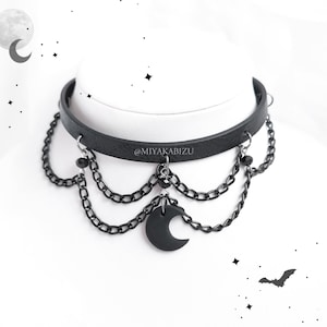 Op de afbeelding: Zwarte leren choker ketting met meerdere kettingen en een halve maan hanger. De choker heeft een zwarte leren band met een zilveren gesp en is versierd met meerdere kettingen en een halve maan hanger. De hanger is gemaakt van zwart metaal en heeft een matte afwerking.