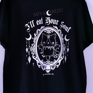 Pode incluir: Camiseta preta com um gr&aacute;fico branco de um gato de desenho animado fofo com asas de morcego em uma moldura decorativa. O texto "I'll eat your soul" est&aacute; acima do gato e "MIYAKABIZU" est&aacute; na parte superior da camiseta.