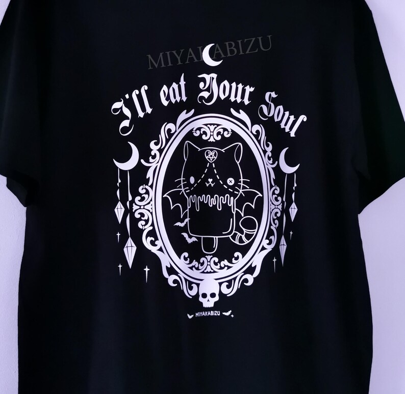 Pode incluir: Camiseta preta com um gr&aacute;fico branco de um gato de desenho animado fofo com asas de morcego em uma moldura decorativa. O texto "I'll eat your soul" est&aacute; acima do gato e "MIYAKABIZU" est&aacute; na parte superior da camiseta.