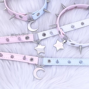May include: Three pastel-coloured collars featuring silver-toned metal spikes, circular eyelets, and dangling charms. Two collars feature silver crescent moon charms, while one features a silver star charm. The text MiyakaBizu is visible.