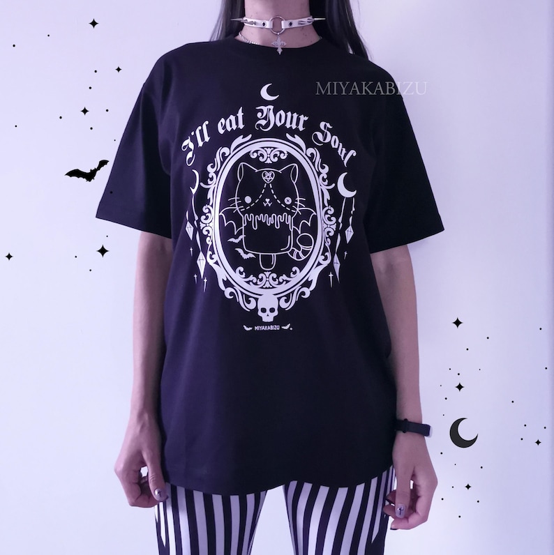 Pode incluir: Camiseta preta com um gr&aacute;fico branco de um gato com asas de morcego em uma moldura de espelho. O texto "I'll eat your soul" est&aacute; escrito em uma fonte g&oacute;tica ao redor do espelho.