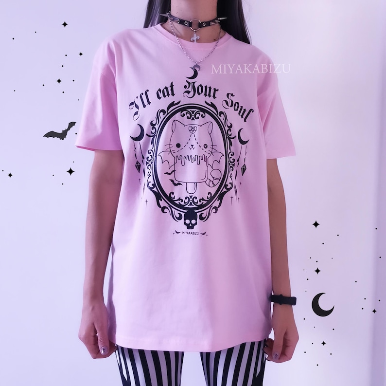 Pode incluir: Camiseta rosa com um gr&aacute;fico preto de um gato com asas em uma moldura. O texto "I'll eat your soul" est&aacute; acima do gato e "MIYAKABIZU" est&aacute; no canto superior direito.