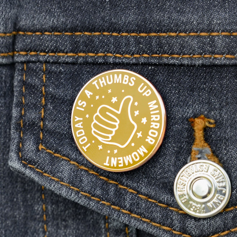 A round enamel pin badge in yellow and gold. The badge shows a thumbs up image and has the words Today is a Thumbs Up Mirror Moment. The badge is pinned to a denim jacket.