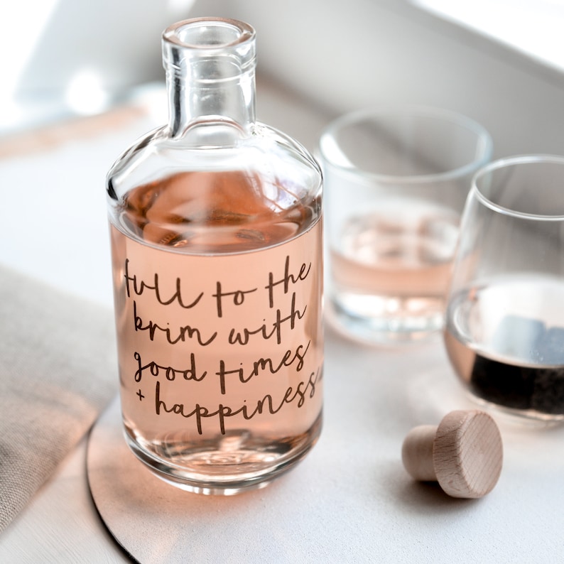 A glass decanter with a wooden stopper next to a glass. The decanter is printed with the words Full to the Brim With Good Times and Happiness.