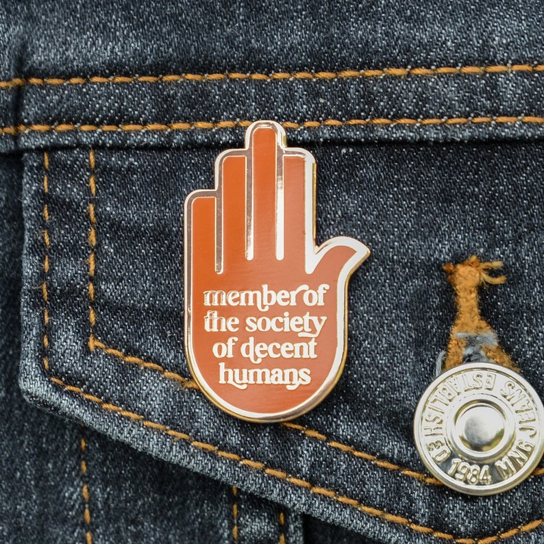 An orange enamel pin badge in the shape of a hand in a pledge style pose.. The text in gold enamel reads Member of the Society of Decent Humans.