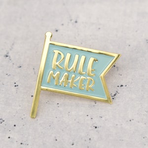 An enamel pin badge in blue and gold. The badge is shaped like a flag and has the text Rule Maker.