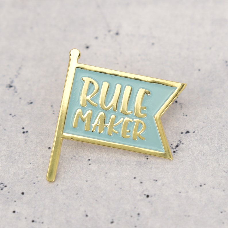 An enamel pin badge in blue and gold. The badge is shaped like a flag and has the text Rule Maker.