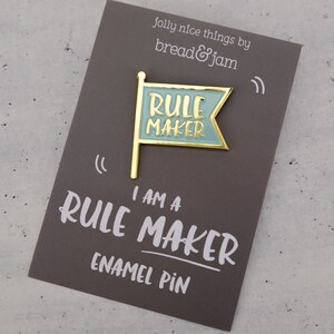 An enamel pin badge in blue and gold. The badge is shaped like a flag and has the text Rule Maker. It is pictured on a backing card and designed by Bread & Jam