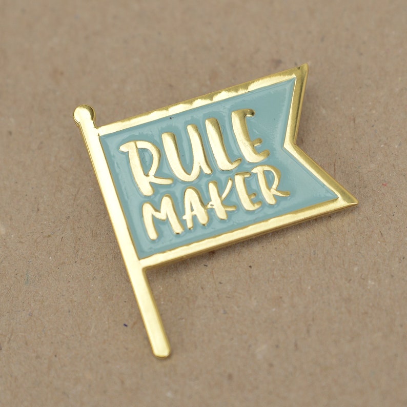 An enamel pin badge in blue and gold. The badge is shaped like a flag and has the text Rule Maker.