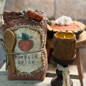 May include: A brown burlap sack with a wooden spoon tied to it, decorated with a pumpkin and the words "Pumpkin Spice". A small candle holder with a lit candle sits next to the sack.