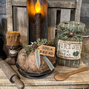 May include: A rustic kitchen scene with a homemade soda bread loaf, a rolling pin, a wooden spoon, and a jar of Irish soda bread. The bread is decorated with greenery and a tag that reads "soda bread".