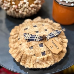 May include: A decorative blueberry pie with a lattice crust, a ruffled edge, and a blue plaid ribbon. A small tag reads "Blueberry pie". The pie is on a dark blue plate, with other decorative items in the background.