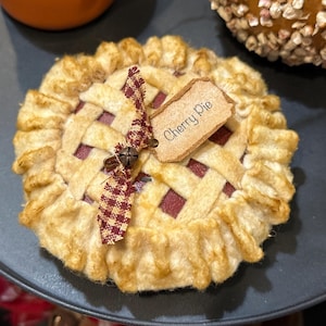 May include: A decorative cherry pie with a lattice crust, displayed on a dark plate. The pie has a red filling and a tag that reads "Cherry Pie". A decorative ribbon and bell accent the pie.