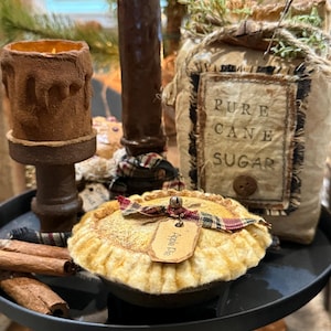 May include: A decorative fabric apple pie with a plaid ribbon and a tag labeled Apple Pie, a fabric bag labeled PURE CANE SUGAR, a brown textured candle holder, and cinnamon sticks arranged on a black tiered tray.