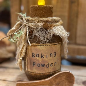 May include: A decorative jar labeled "Baking Powder" with a wooden spoon. The jar has a rustic design with a burlap and net covering, and a small candle on top. The jar is brown with a cream-colored label.