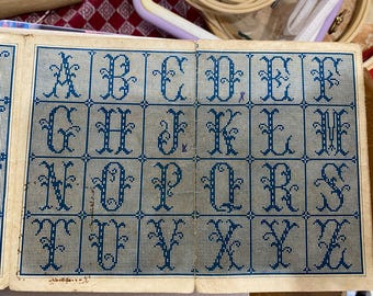 Può includere: Un grafico di alfabeto vintage per punto croce con lettere blu su sfondo bianco. Le lettere sono in un carattere decorativo con fioriture. Il grafico è stampato su carta color crema.