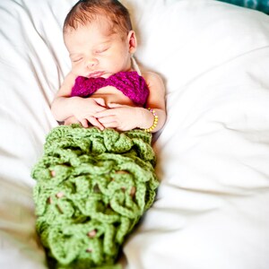 May include: A sleeping baby wearing a green crocheted mermaid tail and a pink crocheted bow. The baby is lying on a white blanket.