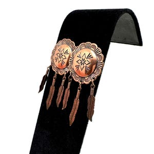 May include: Copper-colored earrings with a circular design and dangling feather charms. The earrings have a detailed, embossed pattern and are displayed on a black velvet stand. The earrings are a vintage style.