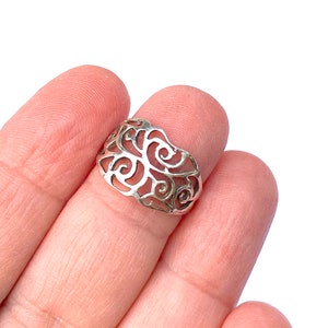 Puede incluir: Un anillo plateado con un dise&ntilde;o calado de patrones arremolinados y en forma de espiral. El anillo se sostiene entre dos dedos, mostrando sus intrincados detalles. El dise&ntilde;o del anillo recuerda formas org&aacute;nicas.