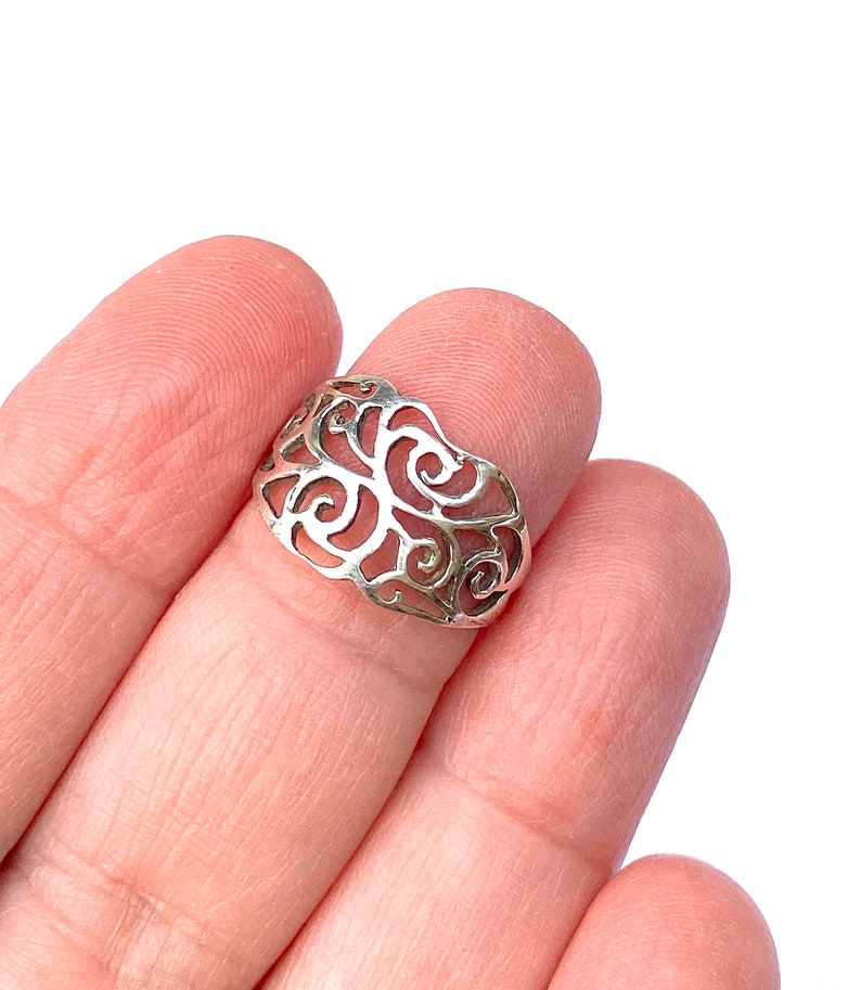 Puede incluir: Un anillo plateado con un dise&ntilde;o calado de patrones arremolinados y en forma de espiral. El anillo se sostiene entre dos dedos, mostrando sus intrincados detalles. El dise&ntilde;o del anillo recuerda formas org&aacute;nicas.