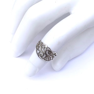 Puede incluir: Un anillo de plata con un dise&ntilde;o calado que presenta patrones sim&eacute;tricos y arremolinados. El anillo se muestra sobre una superficie blanca, mostrando sus intrincados detalles y artesan&iacute;a. El dise&ntilde;o del anillo recuerda motivos florales o de vid.