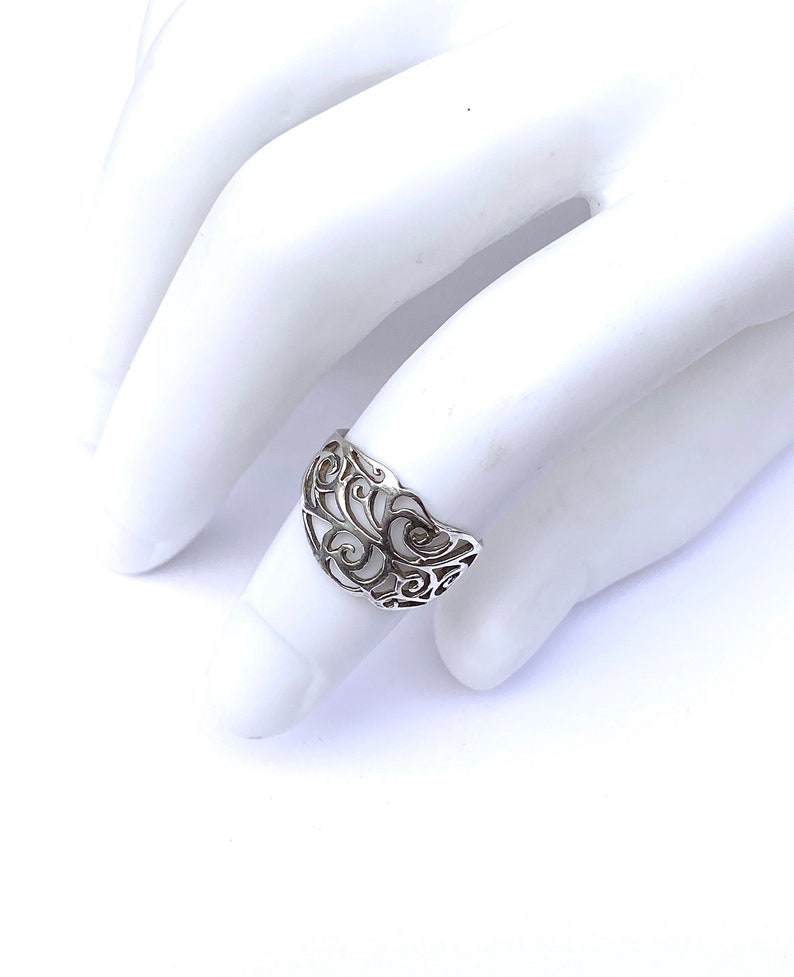 Puede incluir: Un anillo de plata con un dise&ntilde;o calado que presenta patrones sim&eacute;tricos y arremolinados. El anillo se muestra sobre una superficie blanca, mostrando sus intrincados detalles y artesan&iacute;a. El dise&ntilde;o del anillo recuerda motivos florales o de vid.