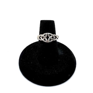 May include: A silver-colored ring with a Celtic knot design is displayed on a black velvet ring holder against a white background. The ring features an interwoven pattern, showcasing a continuous knot design. The ring is a piece of jewelry.