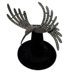 Peut inclure: Broche en forme de colibri argenté, aux ailes déployées, ornée de petites pierres scintillantes. L'oiseau a un long bec incurvé et un petit accent rouge. La broche est présentée sur un présentoir en velours noir sur fond blanc.
