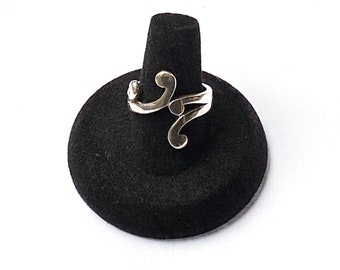 Puede incluir: Un anillo plateado con un diseño abstracto y fluido. El anillo presenta una serie de líneas curvas y espirales, creando un aspecto único. El anillo se muestra sobre un soporte negro para anillos sobre un fondo blanco.