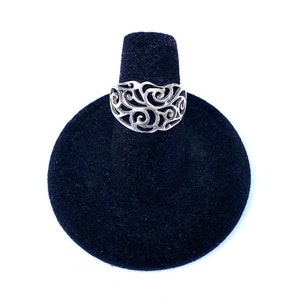 May include: A silver-colored ring with an openwork design featuring swirling patterns. The ring is displayed on a black velvet ring holder against a white background. The ring's intricate details are highlighted.