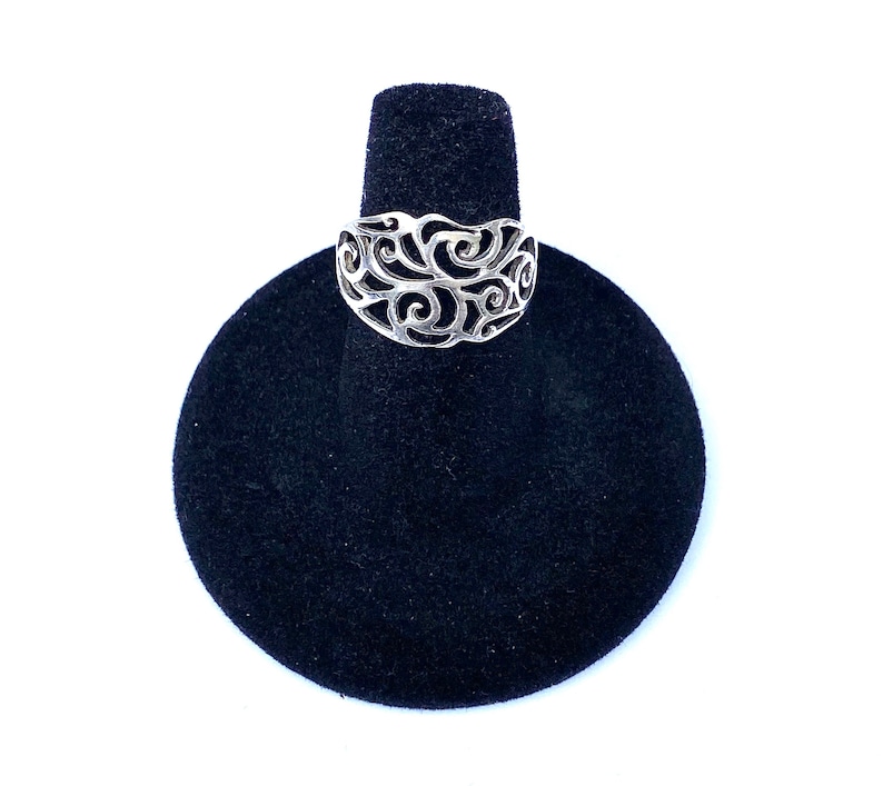 Puede incluir: Un anillo plateado con un dise&ntilde;o calado de patrones arremolinados. El anillo se exhibe sobre un soporte de anillo de terciopelo negro sobre un fondo blanco. Se destacan los intrincados detalles del anillo.