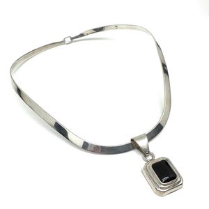 May include: A silver necklace featuring a rectangular black onyx pendant. The necklace has a sleek, curved band that fastens at the back. The pendant is set within a silver frame, creating a striking contrast.