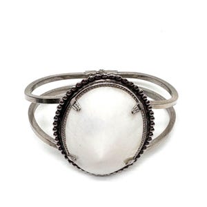 May include: Silver-toned cuff bracelet featuring an oval, white stone centerpiece. The stone is framed by a dark, beaded border and accented with small, decorative elements. The bracelet has a hinged design and a double-banded structure.