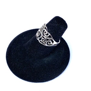 Puede incluir: Un anillo de plata con un dise&ntilde;o calado con patrones arremolinados. El anillo se exhibe en un soporte de anillo de terciopelo negro sobre un fondo blanco. Se destacan los intrincados detalles del anillo.