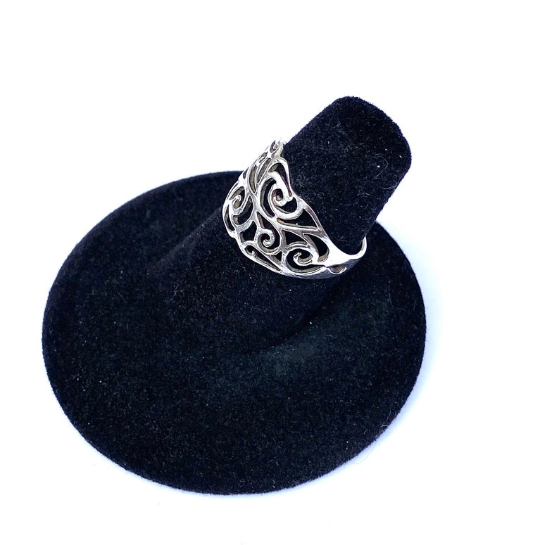Puede incluir: Un anillo de plata con un dise&ntilde;o calado con patrones arremolinados. El anillo se exhibe en un soporte de anillo de terciopelo negro sobre un fondo blanco. Se destacan los intrincados detalles del anillo.