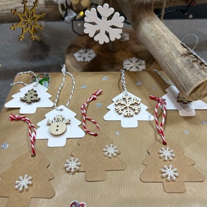 May include: A set of eight brown paper gift tags shaped like Christmas trees. Each tag has a white snowflake or snowman design and a red and white string.